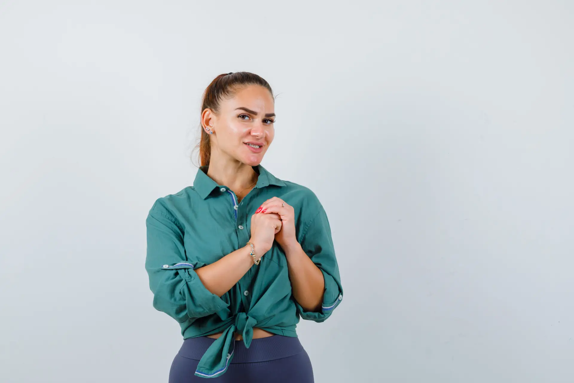 young lady holding clasped hands over chest in green shirt and looking merry , front view.